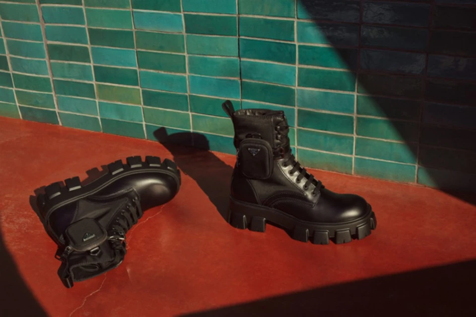 Fashionable black leather combat boots with a pocket detail displayed on a terracotta surface against a textured green tile wall.