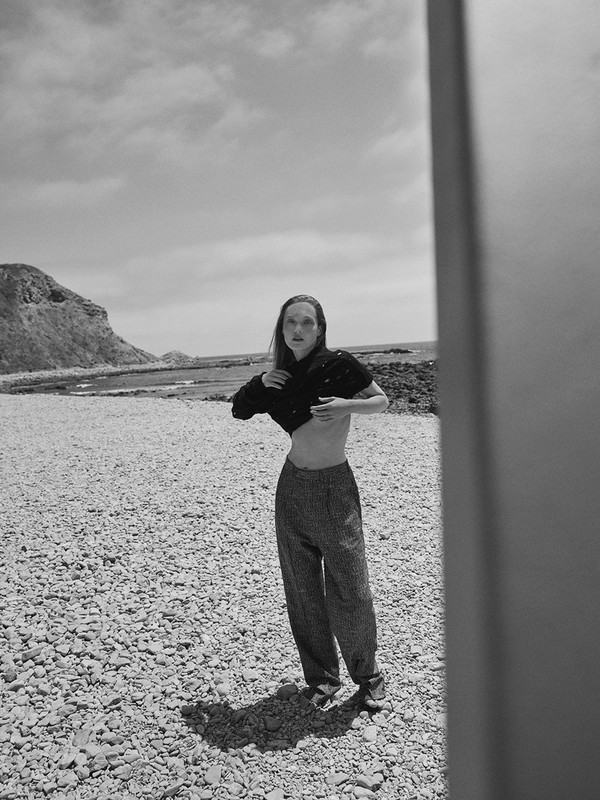 Black and white portrait of a woman posing on a rocky beach in a black top and pants, under a partly cloudy sky with cliffs in background.