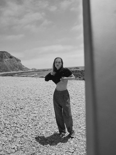 Black and white portrait of a woman posing on a rocky beach in a black top and pants, under a partly cloudy sky with cliffs in background.