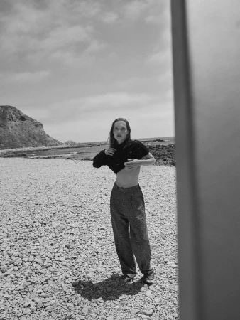 Black and white portrait of a woman posing on a rocky beach in a black top and pants, under a partly cloudy sky with cliffs in background.