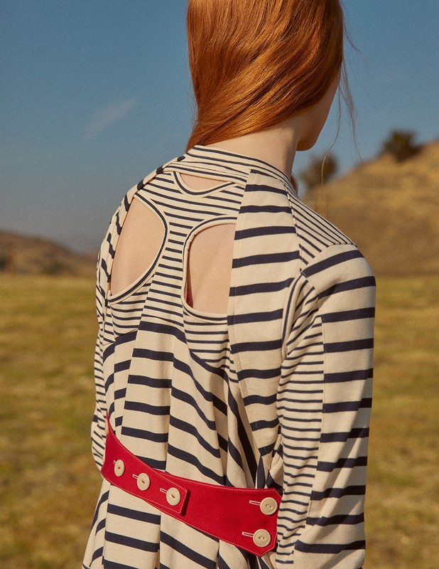 Back view of a woman with red hair wearing a striped cut-out dress and red belt, posing in a grassy field under a clear blue sky.