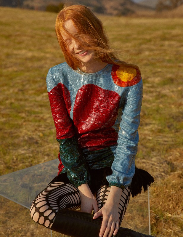 A red-haired woman in an embellished top smiles in a golden field with distant hills, shot in natural light on a sunny day.