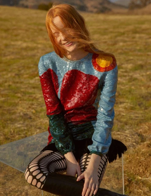 A red-haired woman in an embellished top smiles in a golden field with distant hills, shot in natural light on a sunny day.