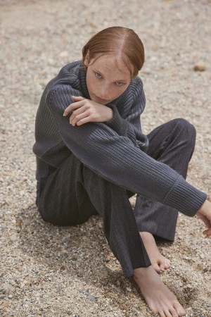 Young woman in a dark grey ribbed sweater and matching trousers sitting barefoot on a pebble beach, photographed by Williams + Hirakawa.