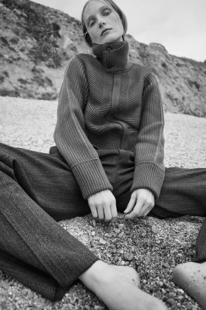 Model seated on a pebble beach wearing a heavy ribbed turtleneck sweater and trousers by photographers Williams + Hirakawa.