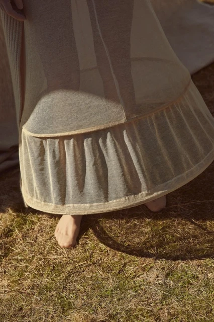 Person standing on grassy field wearing a long dress with ruffle details during daytime, illuminated by sunlight.