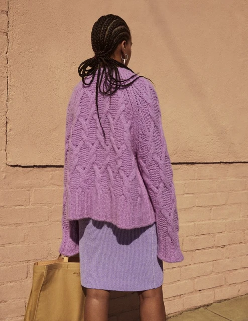 A woman from behind wearing a purple knit sweater and skirt stands against a peach wall, holding a tote bag in a bright outdoor setting.
