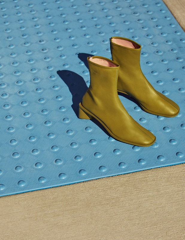 Pair of green ankle boots with a block heel, displayed on a blue textured surface with a concrete background, illuminated by sunlight.