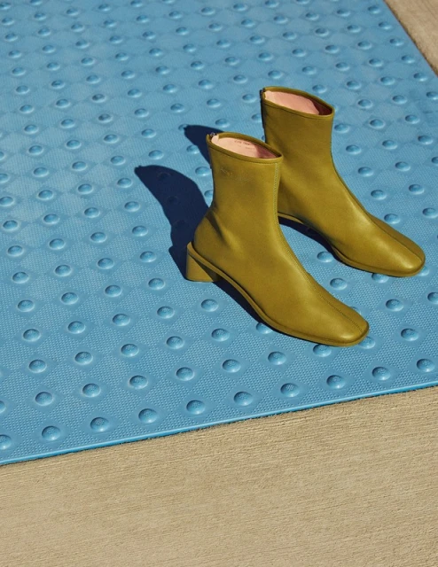 Pair of green ankle boots with a block heel, displayed on a blue textured surface with a concrete background, illuminated by sunlight.