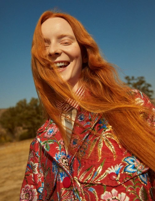 Portrait of a cheerful woman with long red hair, wearing a colorful patterned jacket, smiling outdoors against a blue sky.