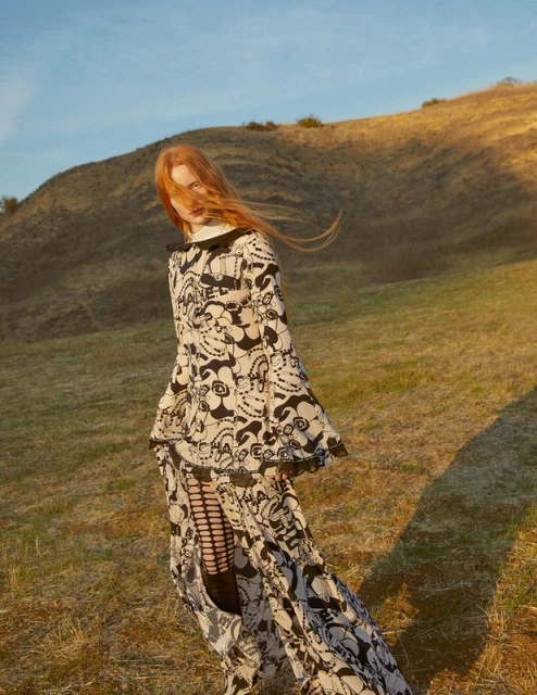A woman in a black-and-white patterned dress stands in a grassy field against a backdrop of rolling hills and a clear blue sky.
