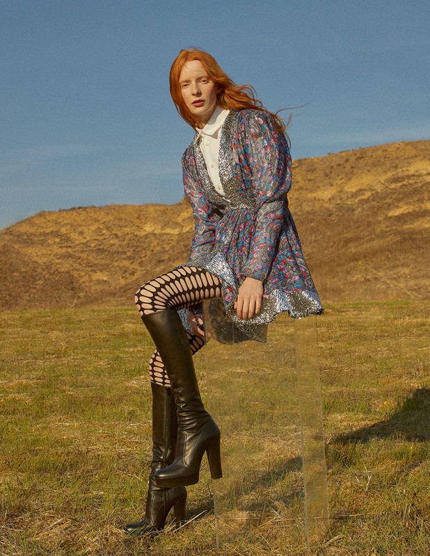 A red-haired woman in a patterned dress and black boots sits on a transparent box outdoors in a grassy field with a brown hill in the background.