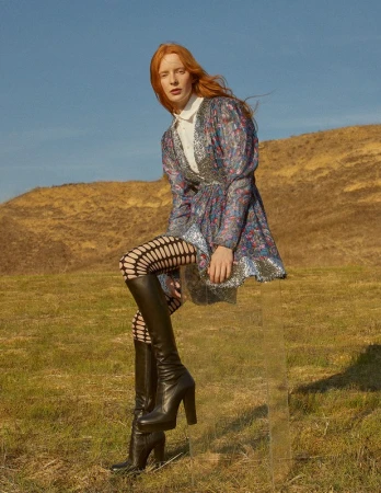A red-haired woman in a patterned dress and black boots sits on a transparent box outdoors in a grassy field with a brown hill in the background.
