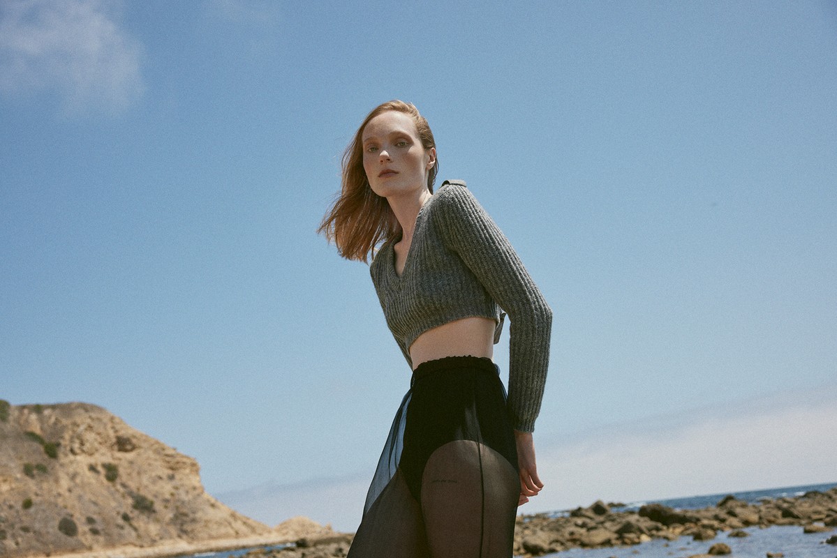A woman in a grey knit crop top poses in front of a blue sky near the beach. The image is taken with a low angle.
