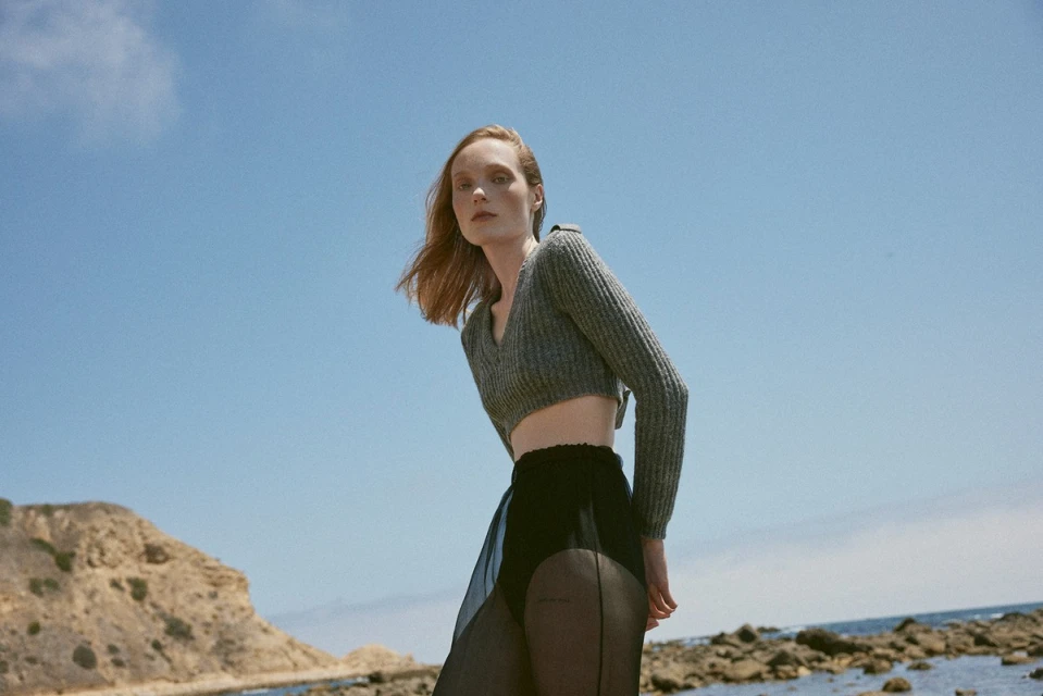 A woman in a grey knit crop top poses in front of a blue sky near the beach. The image is taken with a low angle.