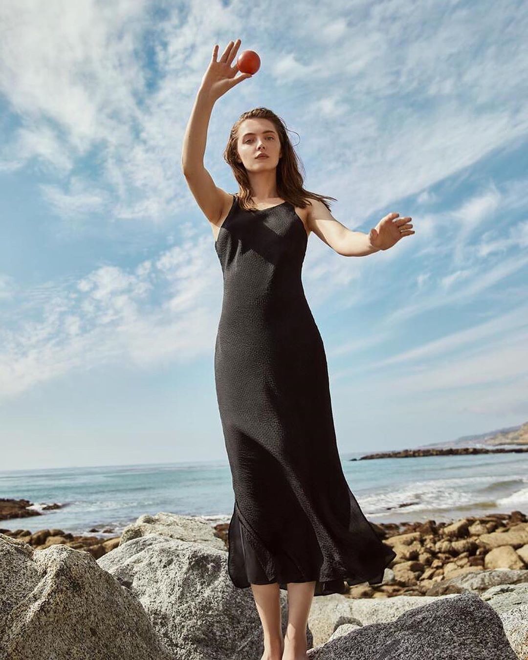 A woman in a black dress stands barefoot on rocks by the ocean, holding a red object, with a cloudy sky above in an outdoor setting.