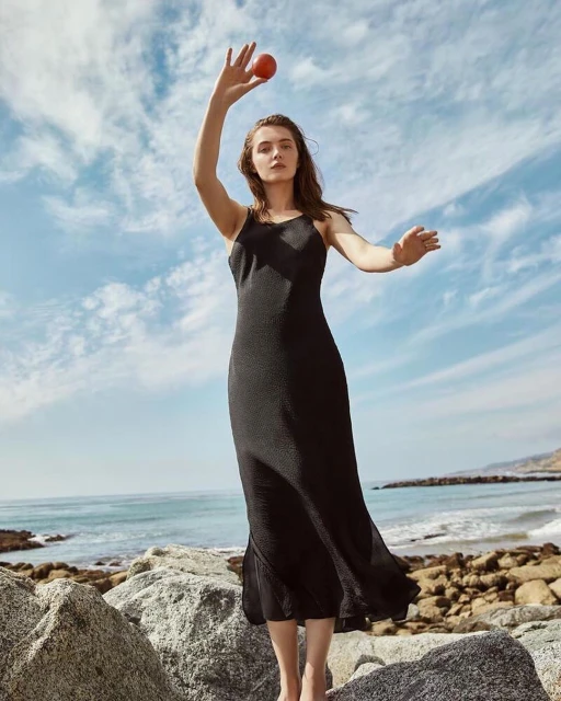 A woman in a black dress stands barefoot on rocks by the ocean, holding a red object, with a cloudy sky above in an outdoor setting.