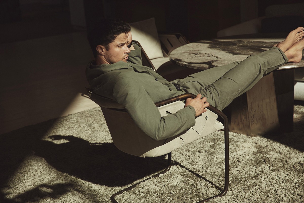 An adult male lounges in a chair indoors with natural light creating strong shadows and highlights on the neutral-toned carpet and furniture.