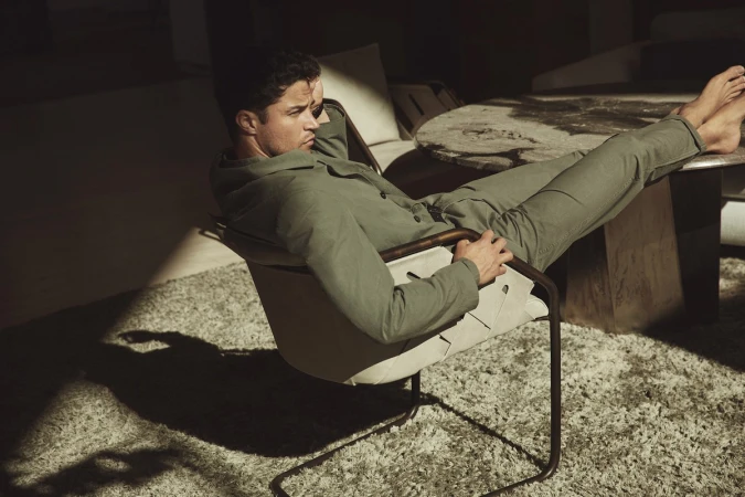 An adult male lounges in a chair indoors with natural light creating strong shadows and highlights on the neutral-toned carpet and furniture.
