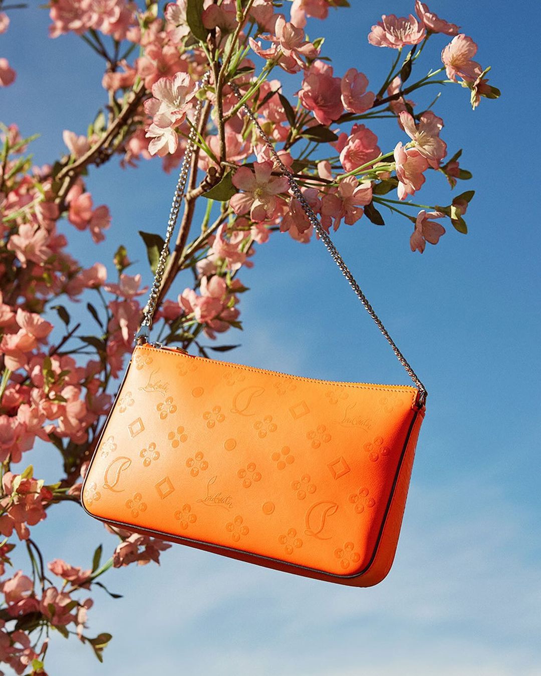 Orange handbag with a silver chain strap is suspended from a flowering tree branch against a bright blue sky in an outdoor setting.