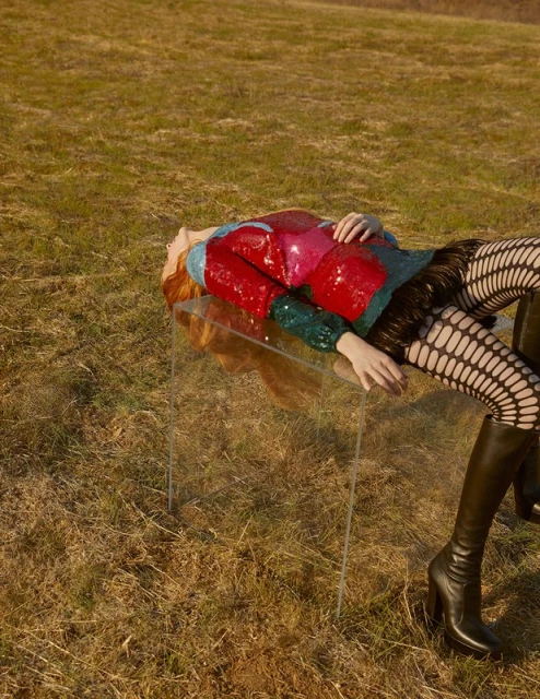 Woman in sequin jacket, fishnet stockings, and boots lying on a clear table outdoors in a field of dry grass on a sunny day.