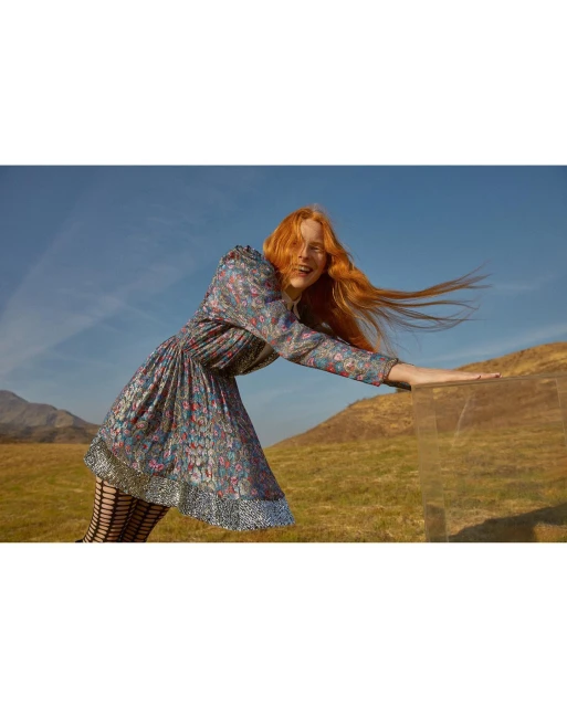 A red-haired woman in a floral dress smiles in a field. The model wears patterned tights with a glass box in the foreground.