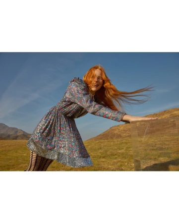 A red-haired woman in a floral dress smiles in a field. The model wears patterned tights with a glass box in the foreground.