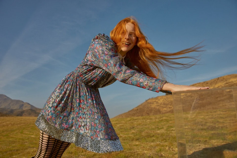 A red-haired woman in a floral dress poses outdoors with mountains in the distance, leaning on a transparent box under a blue sky.