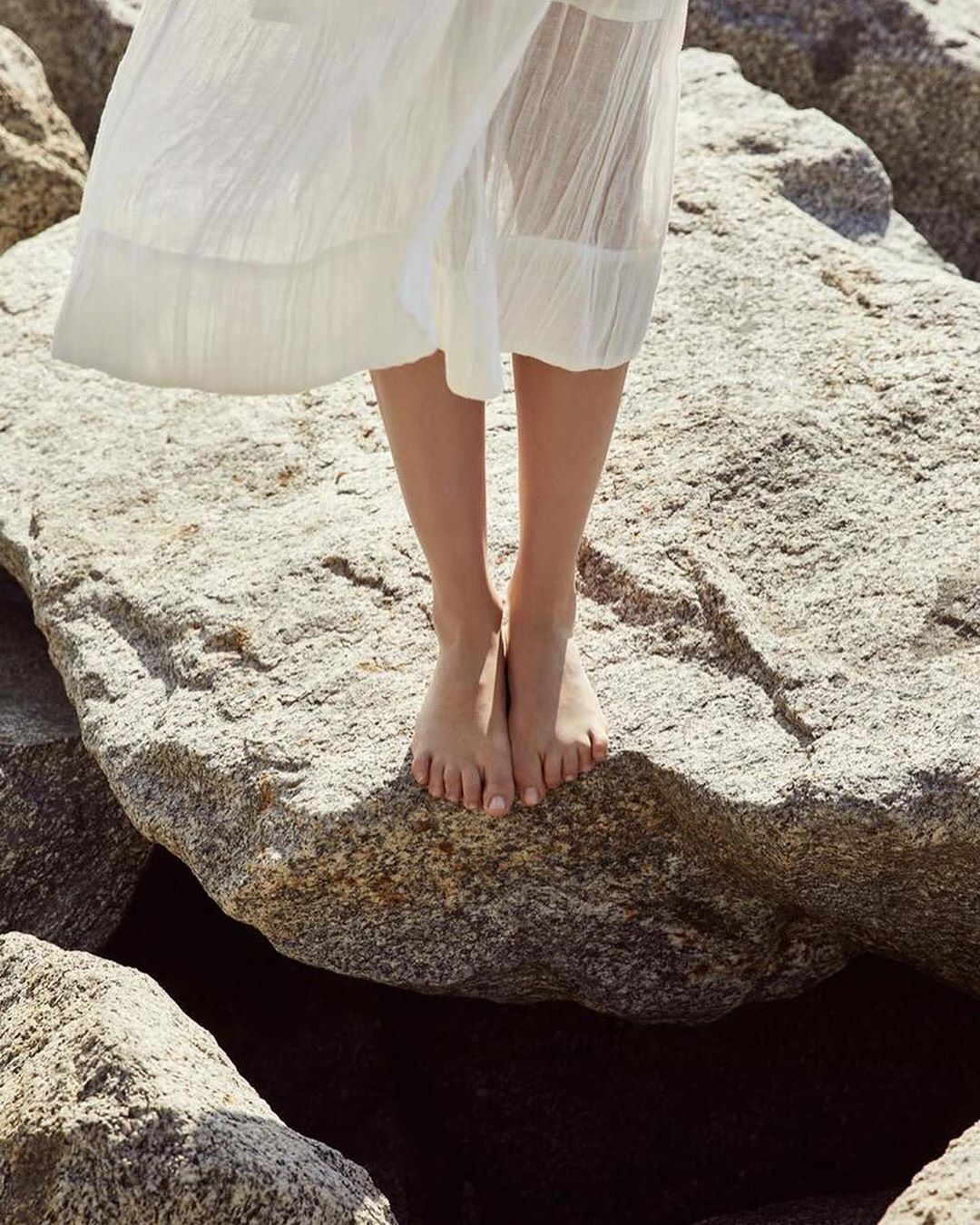 Woman in a white dress stands barefoot on large rocks by the water, showcasing a minimalist style in a natural setting on a sunny day.