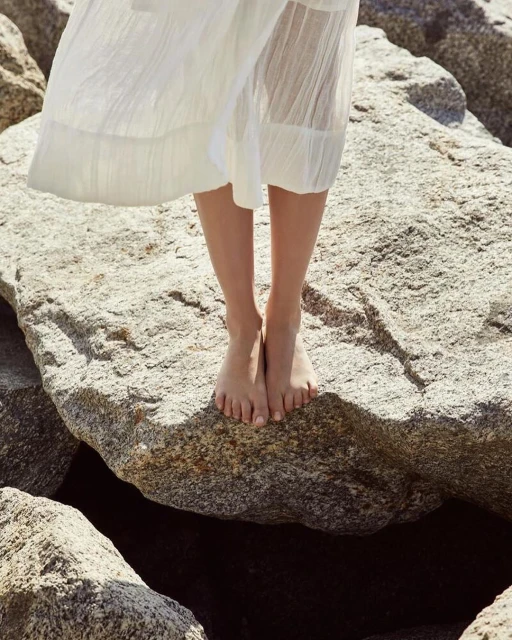 Woman in a white dress stands barefoot on large rocks by the water, showcasing a minimalist style in a natural setting on a sunny day.