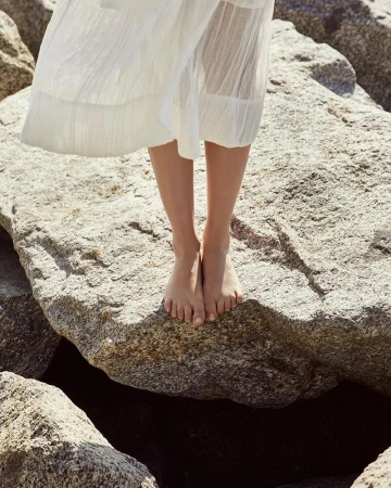 Woman in a white dress stands barefoot on large rocks by the water, showcasing a minimalist style in a natural setting on a sunny day.