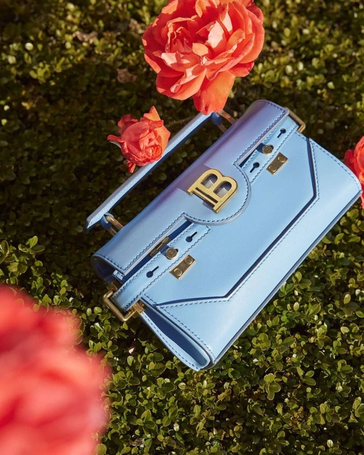 Light blue Balmain handbag with gold hardware and logo on green foliage with peach colored roses in outdoor fashion setting.