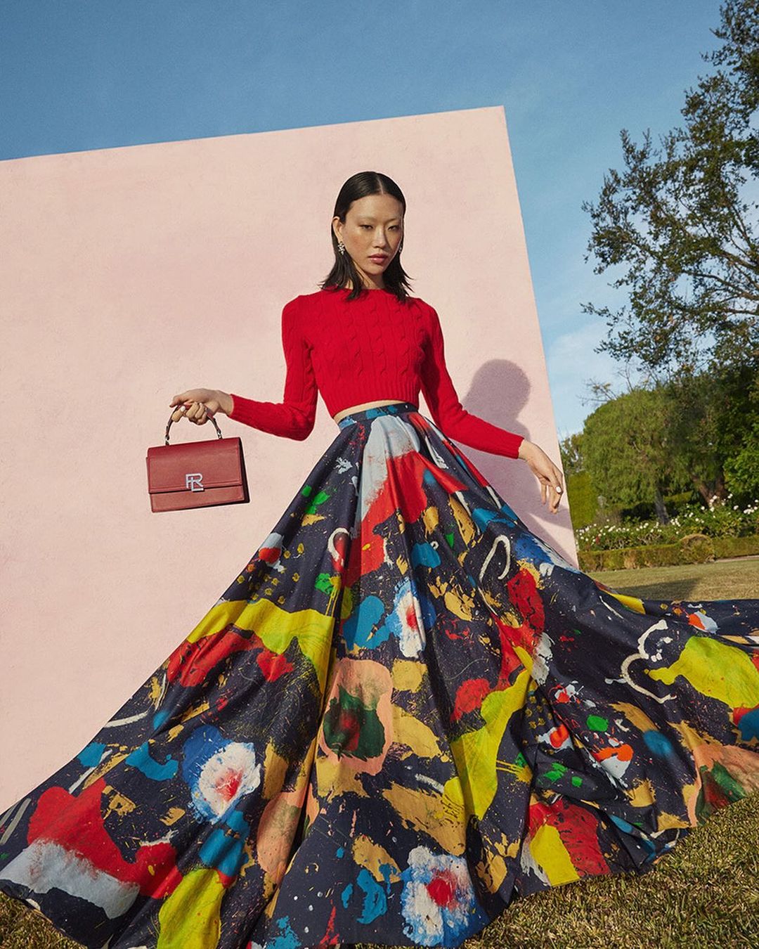Model in red crop top and printed maxi skirt holding a handbag against a pink wall by photographers Williams + Hirakawa.