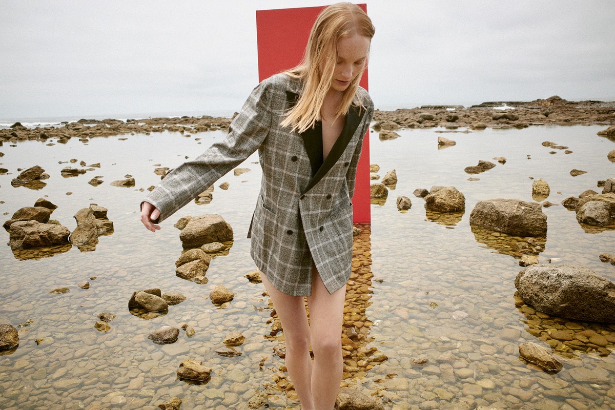 A fair-skinned woman in a grey plaid blazer walks through shallow water on a rocky beach with a red vertical panel in background.
