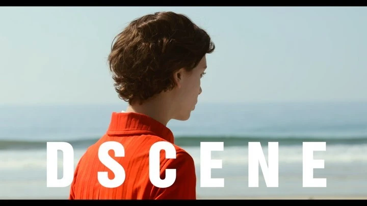Adult with short curly hair stands near ocean, wearing an orange ribbed top, facing away from the camera in a medium shot.