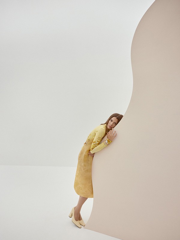 A woman in a yellow dress and heels leans against a light pink curved wall in a neutral studio environment, in a fashion editorial style.