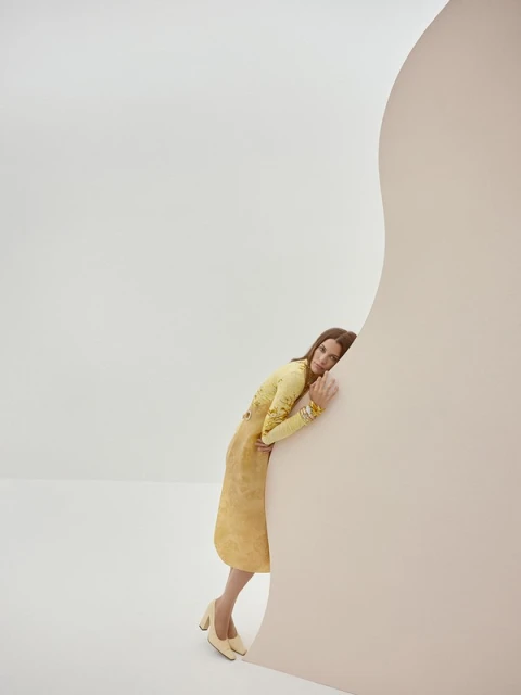 A woman in a yellow dress and heels leans against a light pink curved wall in a neutral studio environment, in a fashion editorial style.