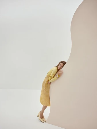 A woman in a yellow dress and heels leans against a light pink curved wall in a neutral studio environment, in a fashion editorial style.
