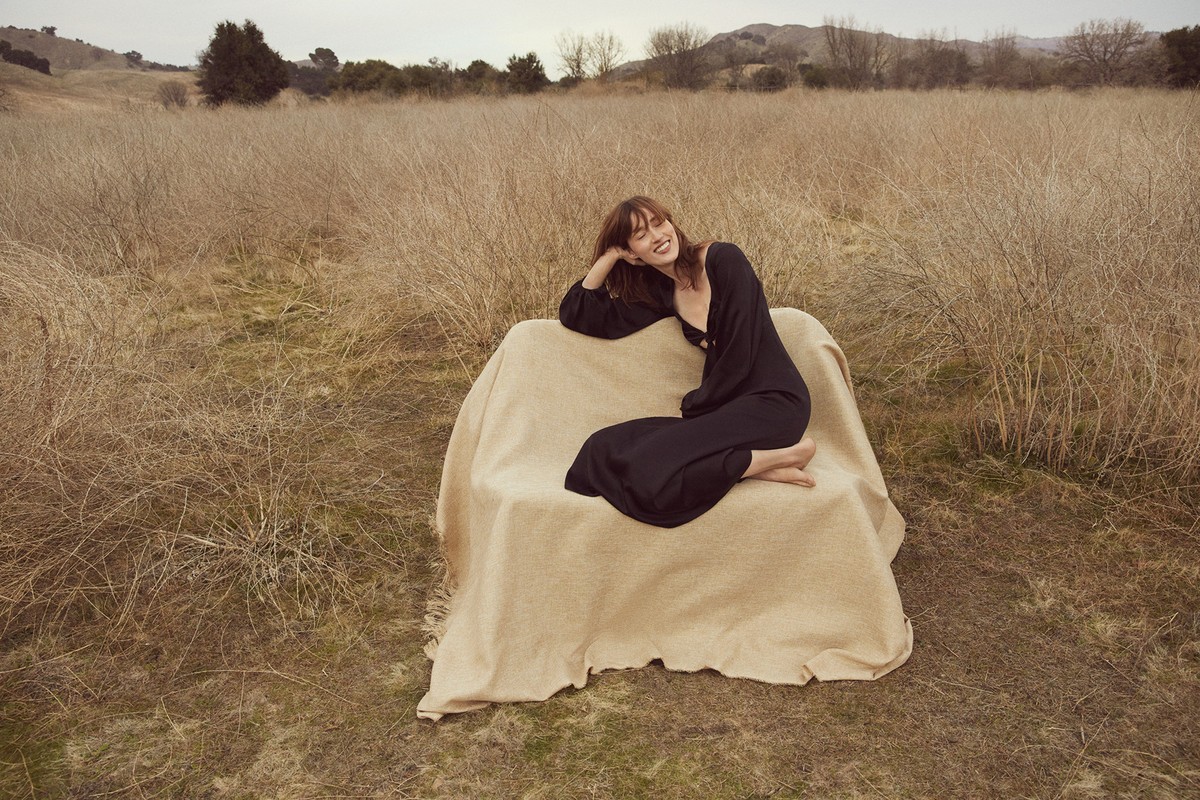 A woman in a black dress sitting on a beige-covered chair in a dry field with rolling hills in the background under an overcast sky.