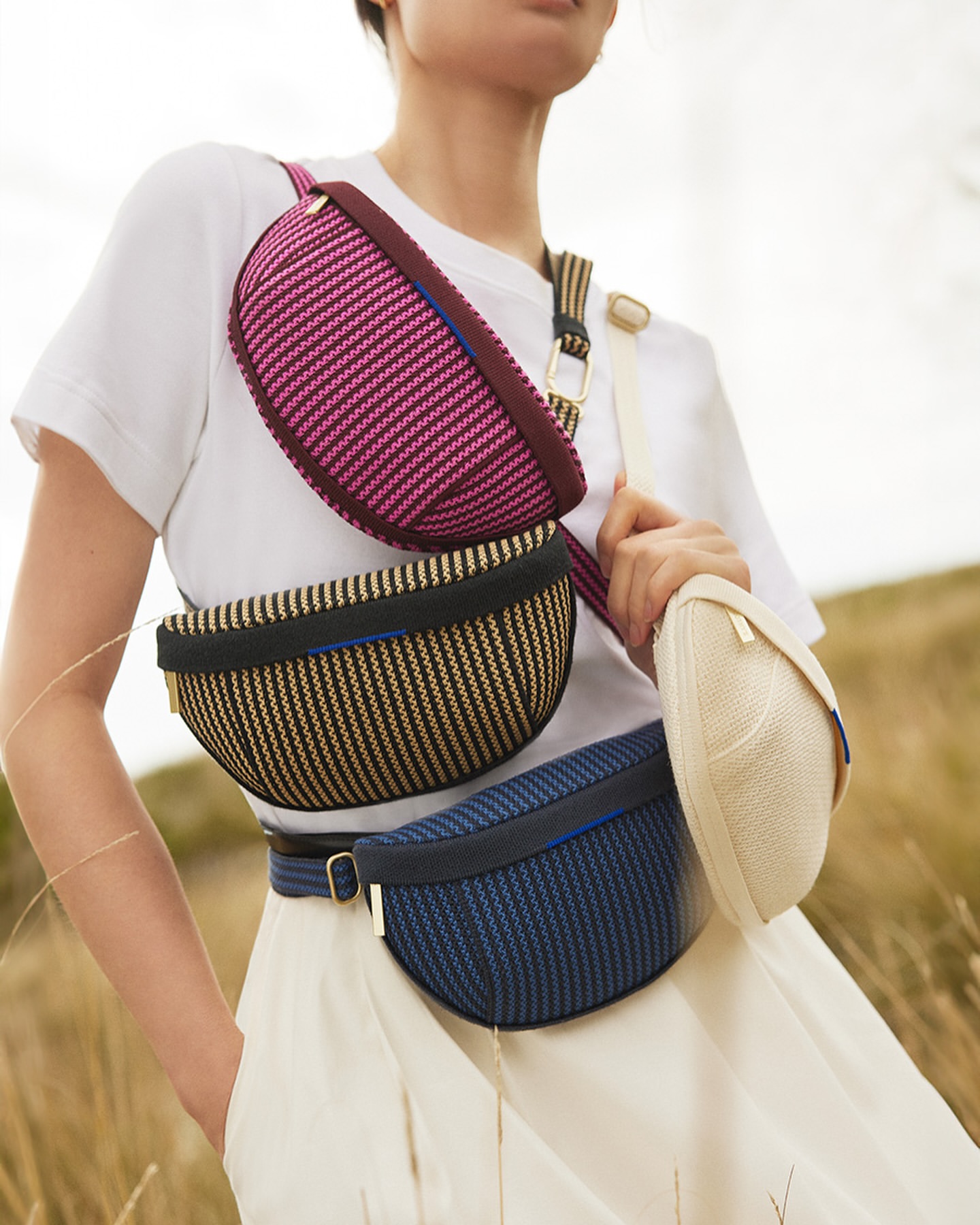 Eye-level shot of a woman wearing a white t-shirt and skirt, displaying multiple colorful woven bags strapped across her chest and waist.