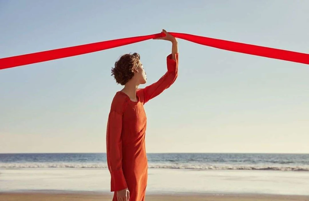 Woman in a red dress on a beach holds a red ribbon against a blue sky and ocean backdrop, creating a striking color contrast.