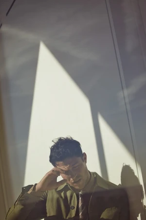 Portrait of a thoughtful adult man in olive green trench coat with hand to forehead in a brightly lit indoor space, with stark shadows.