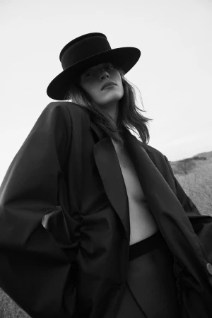 Monochrome portrait of a model wearing a wide-brimmed black hat and oversized dark blazer against an outdoor landscape background.