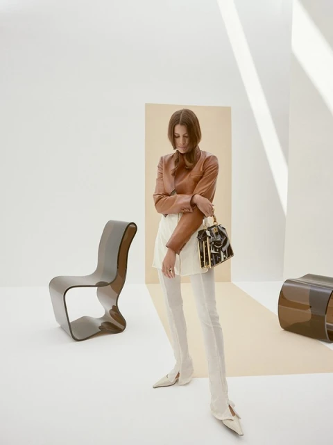 Fashionable adult female in stylish brown top, white skirt, and long boots posing with a luxury purse in bright studio with chairs.