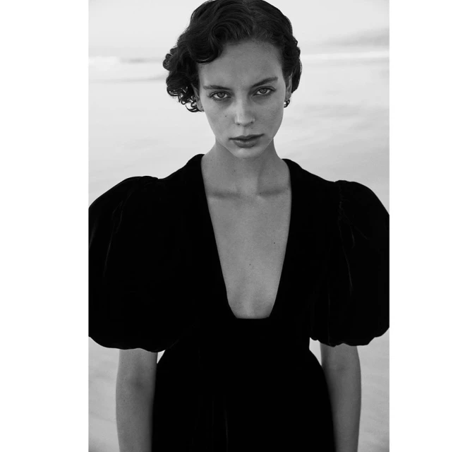Monochrome portrait of a woman in a black velvet dress with puffed sleeves, gazing at the camera, against a blurred light background.