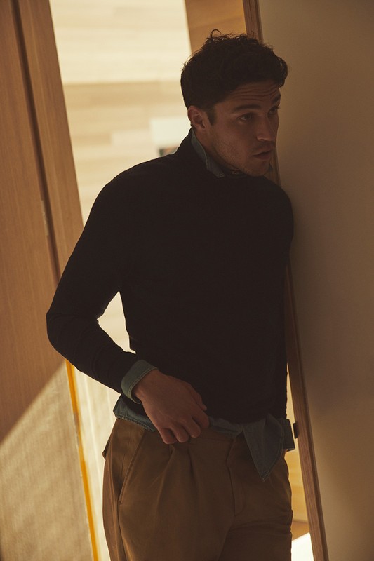 Portrait of a handsome man with dark curly hair, wearing a dark sweater and khaki pants, standing in an indoor doorway, looking to the side.