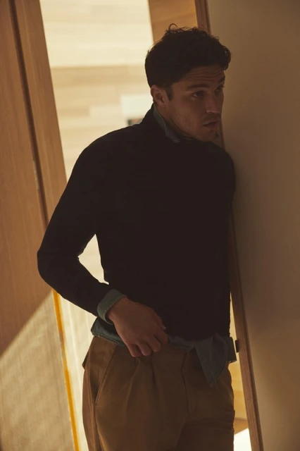 Portrait of a handsome man with dark curly hair, wearing a dark sweater and khaki pants, standing in an indoor doorway, looking to the side.