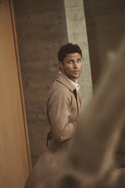 Stylish adult male poses in a studio wearing a brown coat, looking away from the camera, with a neutral expression and modern environment.