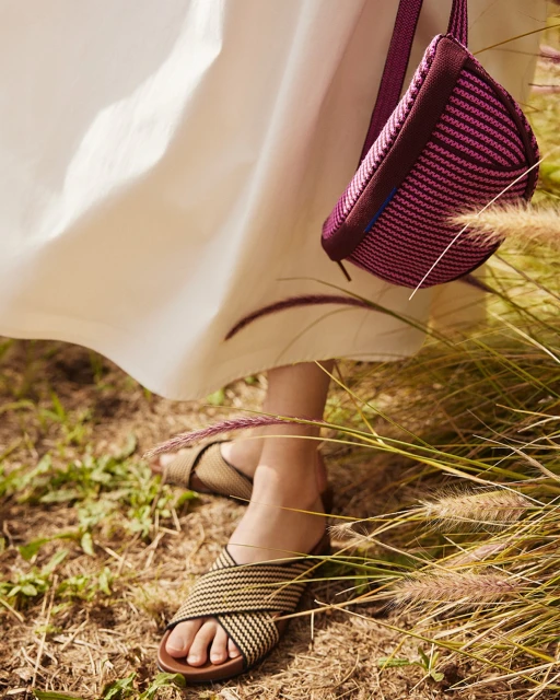 Close-up of woman wearing woven slide sandals with a magenta cross-body bag and long white dress in a field of tall grass.