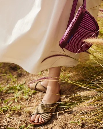 Close-up of woman wearing woven slide sandals with a magenta cross-body bag and long white dress in a field of tall grass.